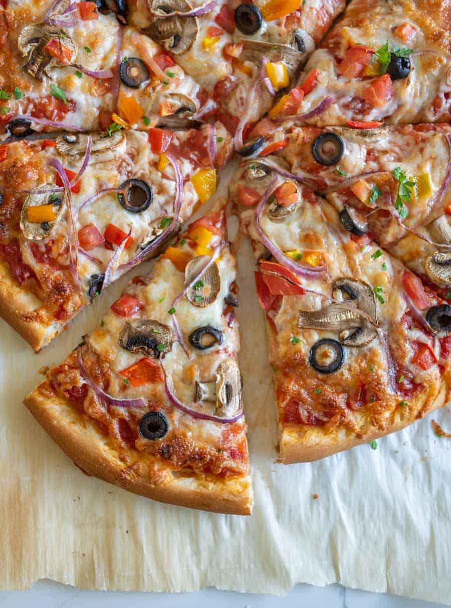 A sliced veggie pizza recipe topped with olives, mushrooms, red onions, tomatoes, and cheese, resting on parchment paper on a light surface.