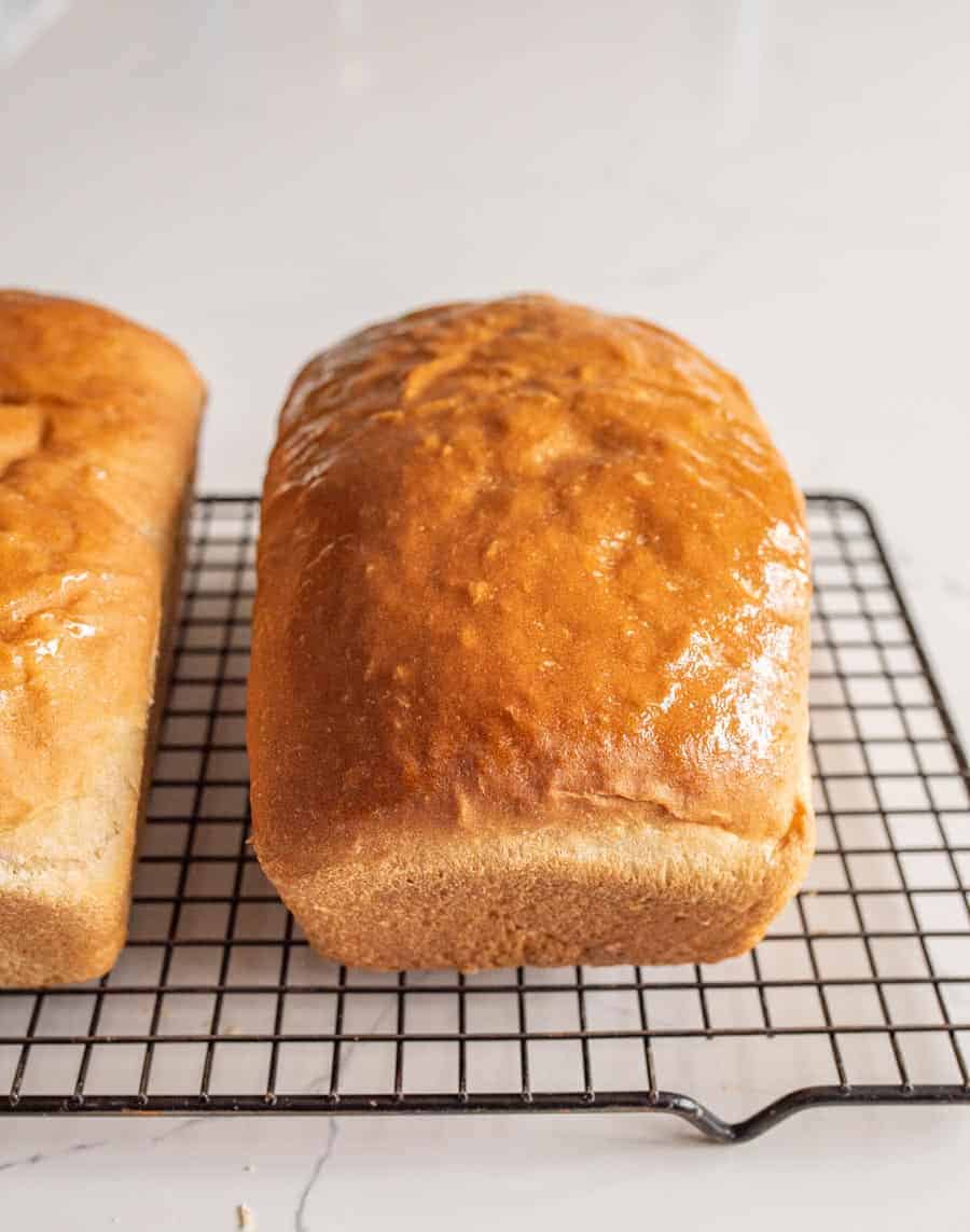 Two freshly baked loaves of golden brown bread, made without eggs, milk, or butter, cool on a black wire rack on a light-colored countertop. Perfect for anyone curious about how to make bread without eggs milk or butter.