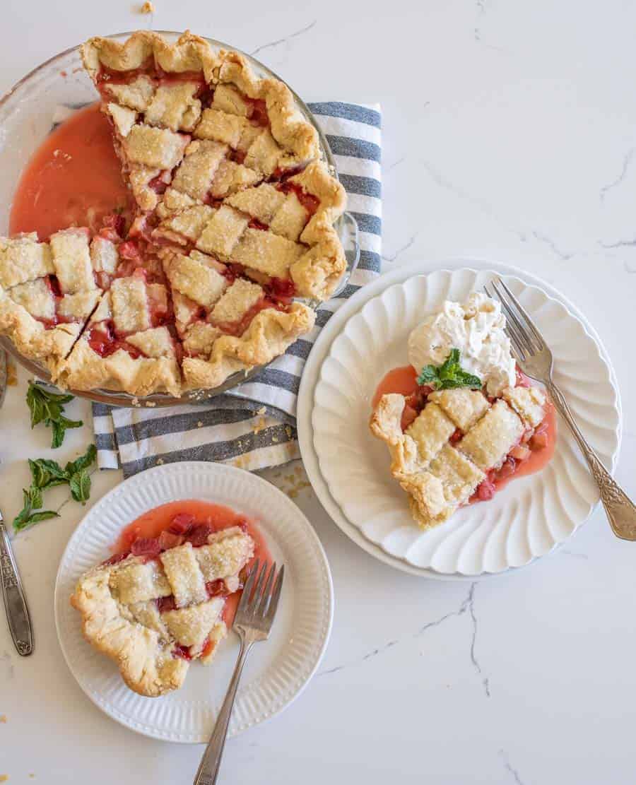 A top view of two slices of strawberry rhubarb pie on white plates next to the rest of the pie in a glass pie pan.