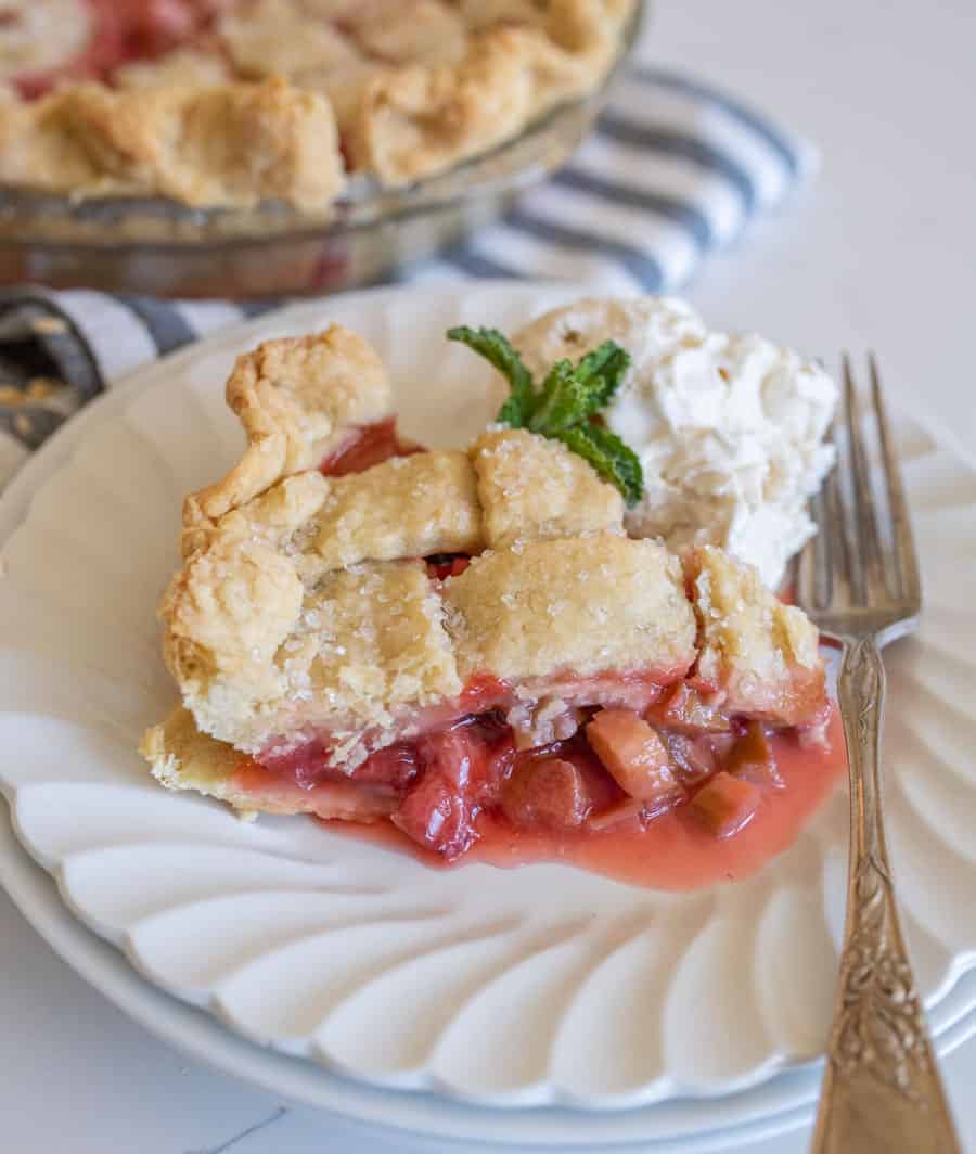 A side view of a slice of strawberry rhubarb pie on a white plate with a scoop of ice cream next to it and a piece of mint.