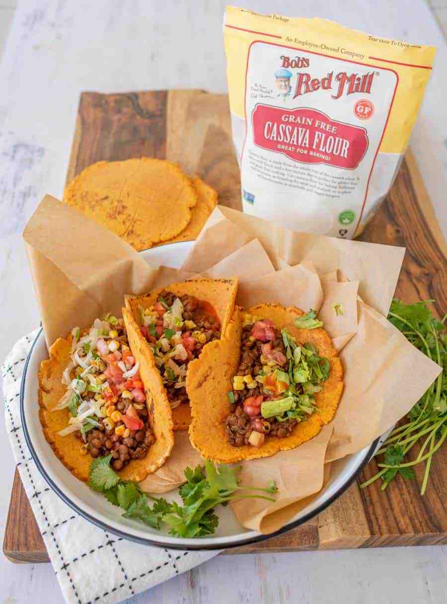 Three sweet potato tortillas with lentil taco filling on parchment paper in a white enamelware pan with a black rim on a wooden board.