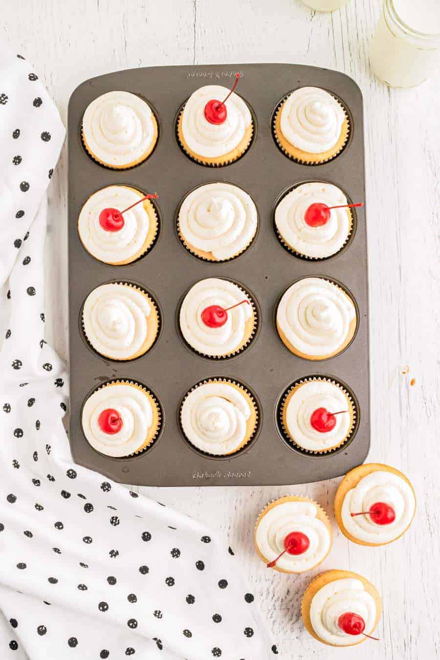 Baked and frosted cherry chip cupcakes in a muffin tin on a white counter.