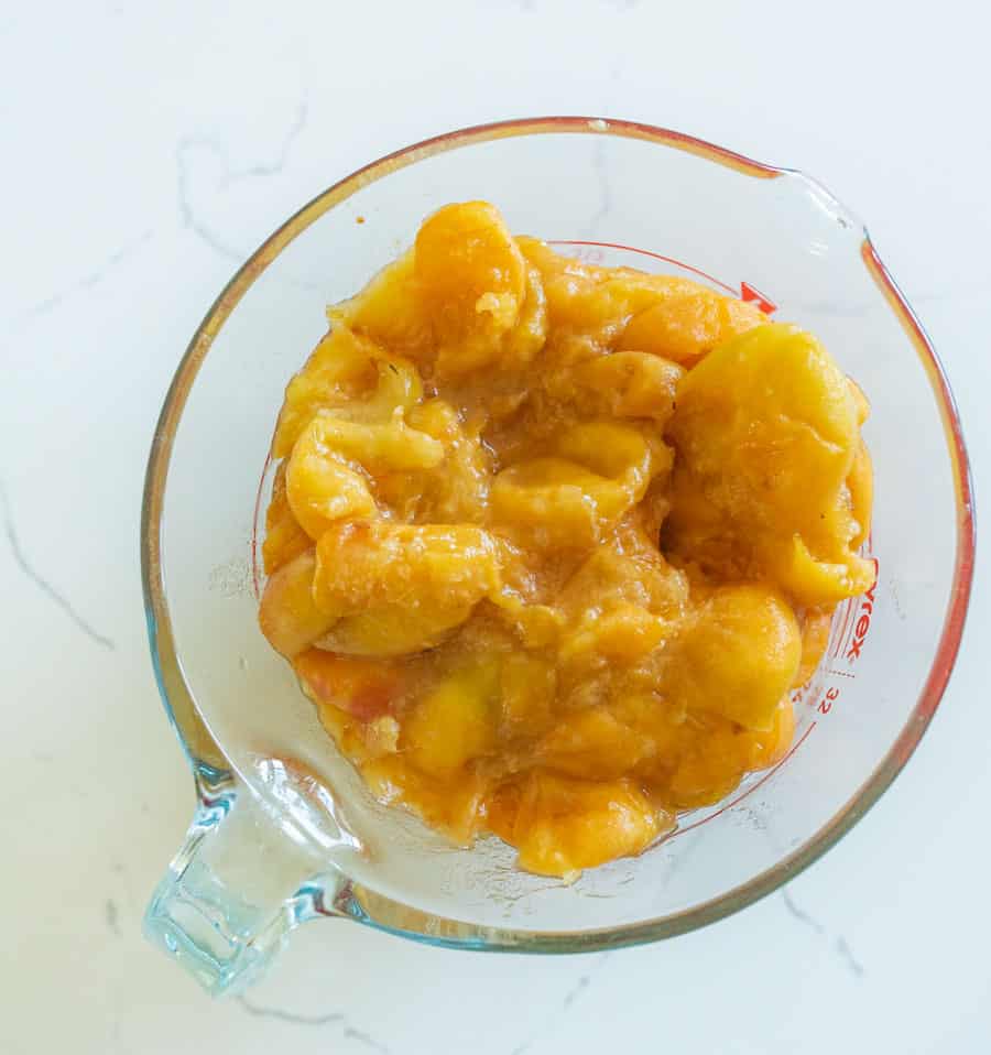 A glass measuring cup filled with mashed peaches sits on a white marble countertop. The peaches appear soft and saucy, with a golden-yellow color—perfect for making easy homemade fruit leather snacks for kids.