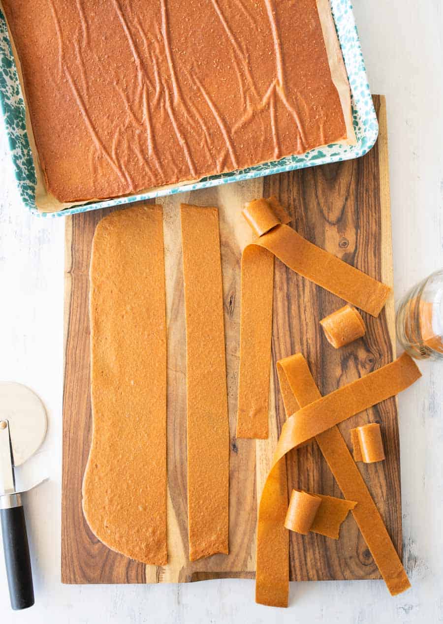 A baking sheet with a large, flat sheet of homemade fruit leather, partially cut into long strips on a wooden board. Some strips are rolled up, and a pizza cutter is nearby.