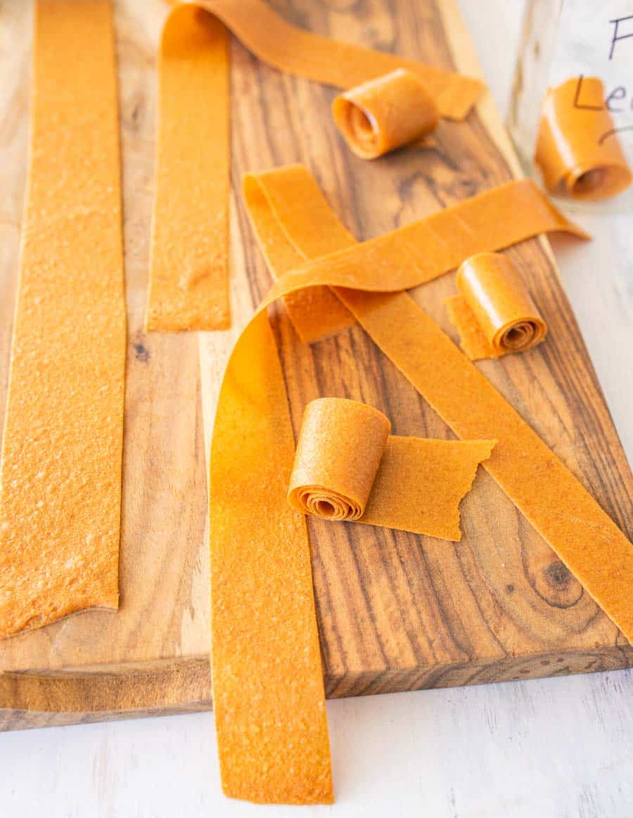Several homemade fruit leather strips and rolls in vibrant orange are displayed on a wooden cutting board, with a light background. Some strips are rolled up, while others are laid flat.