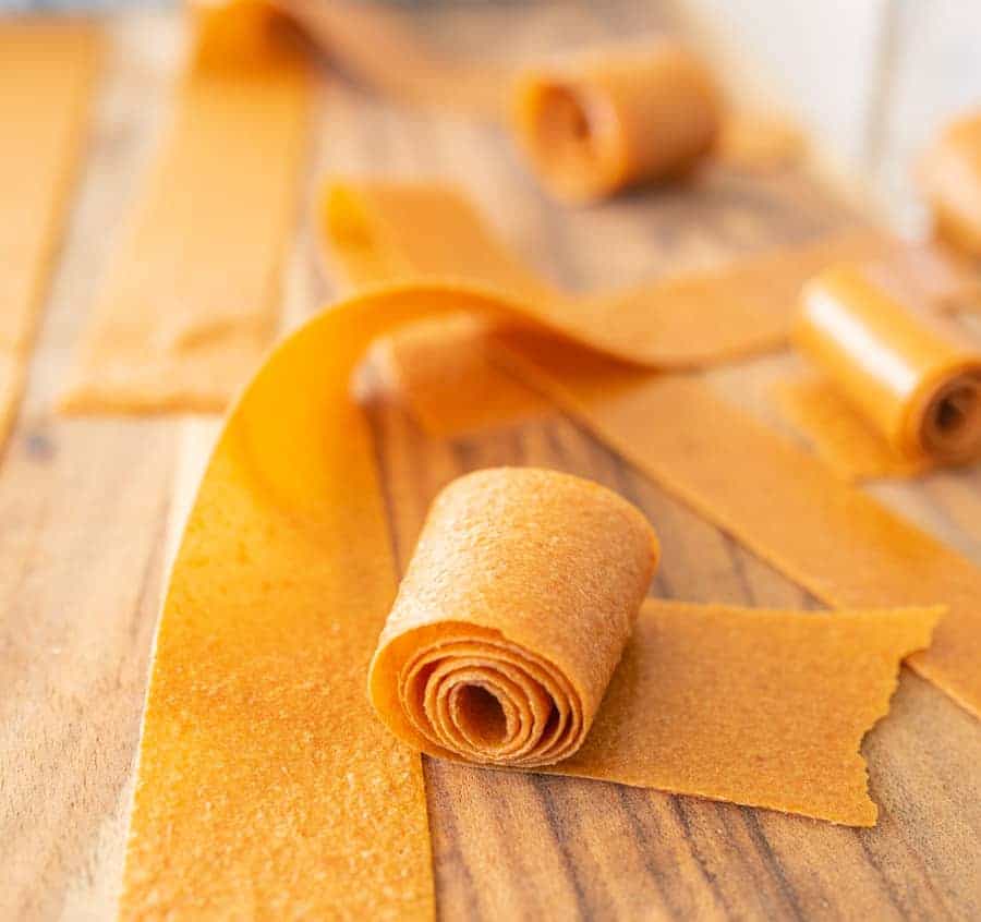 Wide strips of homemade fruit leather are rolled up and laid out on a wooden surface. Some pieces remain flat, while others are partially or fully rolled.