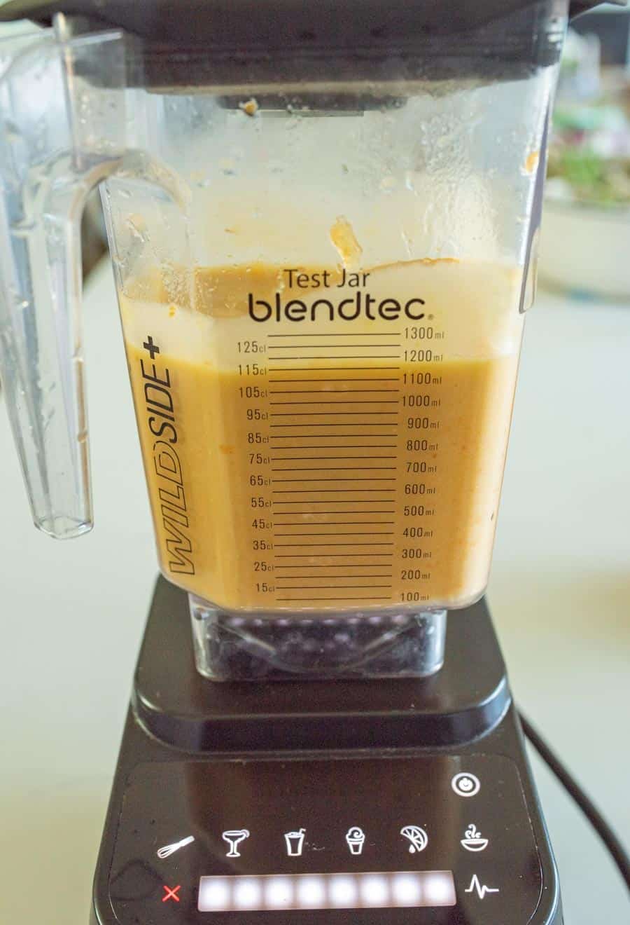 A Blendtec blender jar filled with a thick orange mixture sits on its base, perfect for making homemade fruit leather snacks for kids. The jar features measurement markings and the label Test Jar, while the control panel lights show it’s powered on.