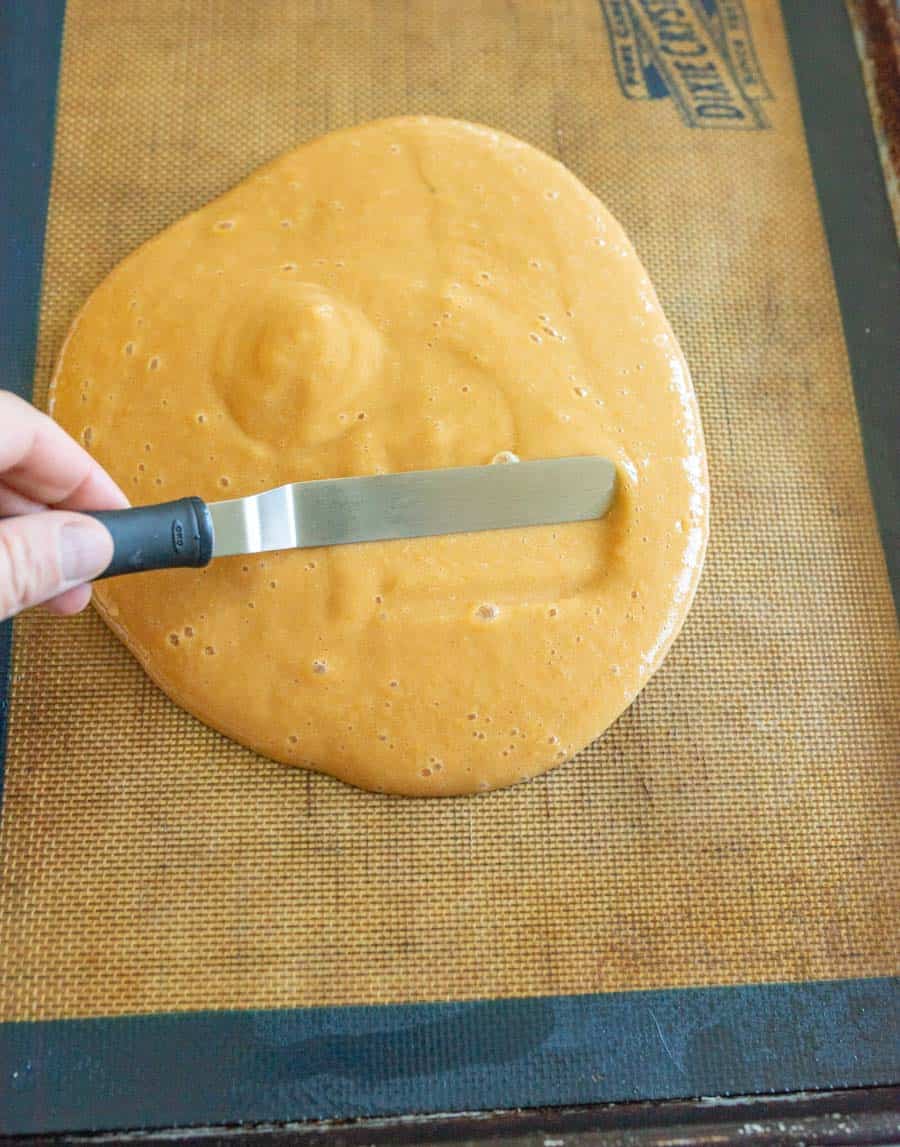 A hand uses an offset spatula to spread a thick, golden caramel mixture—similar in texture to homemade fruit leather—on a silicone baking mat placed on a baking sheet.