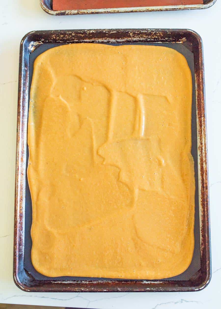 A baking sheet covered with smooth, light brown homemade fruit leather mixture is spread evenly and ready for baking. The well-used pan’s worn edges hint at many batches before.