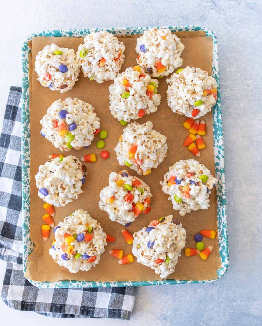 A dozen Halloween popcorn balls with candy corn and M&Ms on an enamel baking sheet.