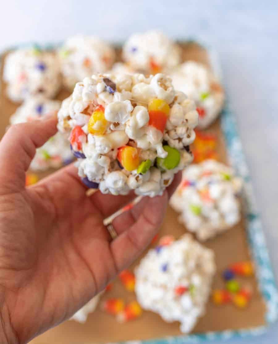 A close-up of a Halloween popcorn ball with M&Ms and candy corn in a hand with additional popcorn balls in the background.