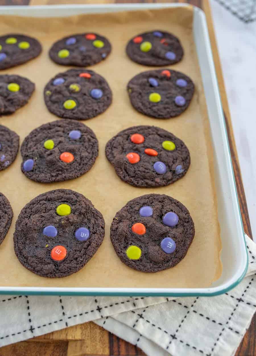 Dark chocolate Halloween M&M cookies after baking on a white enamelware baking sheet lined with parchment paper.