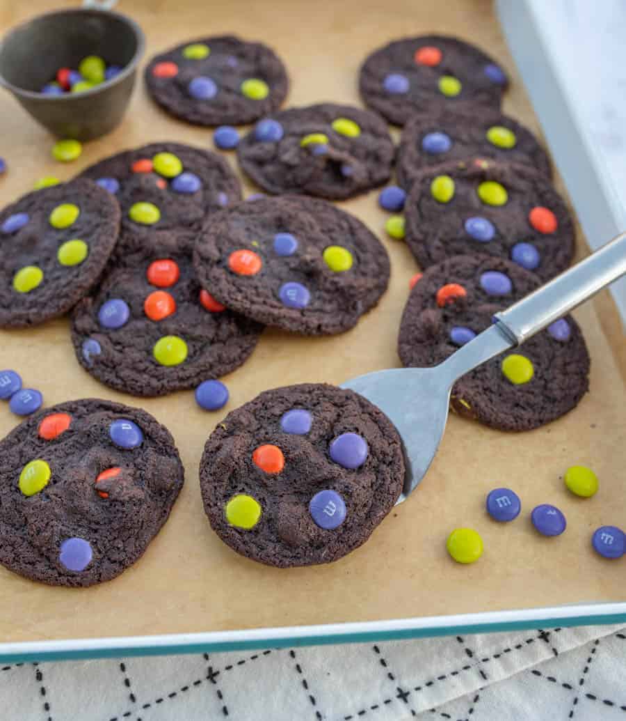 Baking sheet filled with dark chocolate halloween M&M cookies and a metal spatula removing one.