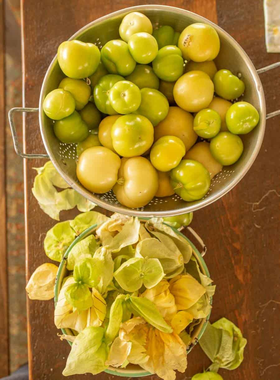 Husked tomatillos in colander with husks in a small bowl.