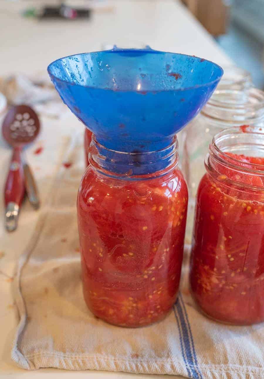 blue funnel in glass jar being filled with tomatoes.