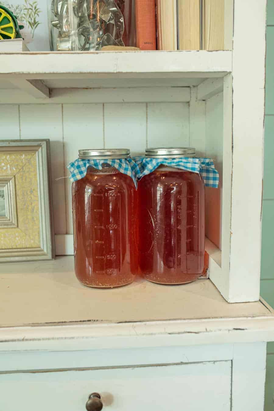 two half gallon mason jar filled with first ferment kombucha and capped with gingham cloth and metal ring sitting on pretty hutch.