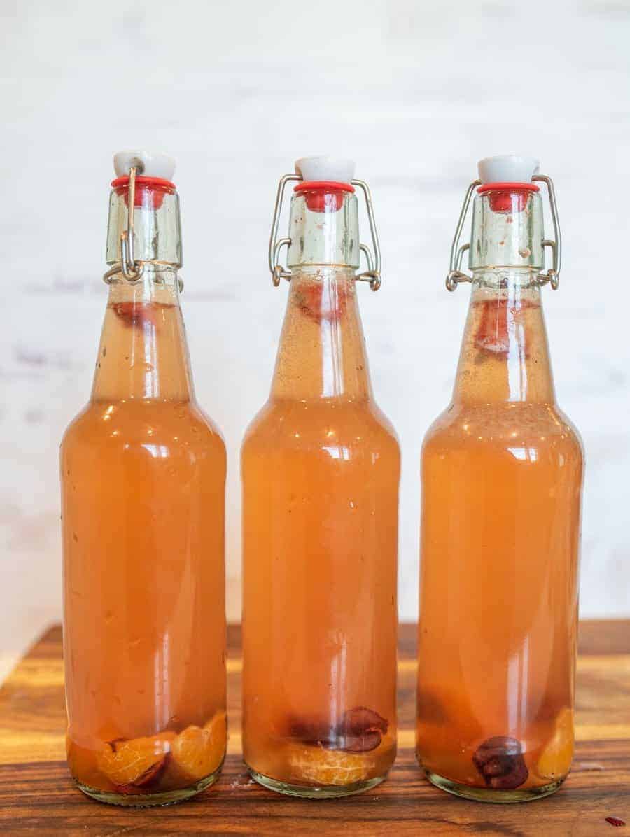 Three clear glass bottles with swing-top lids are filled with a light orange, fizzy homemade kombucha. Pieces of fruit rest at the bottom of each bottle. The bottles sit on a wooden surface against a light background.