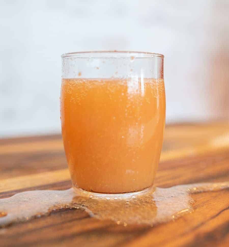 A glass filled with homemade kombucha sits on a wooden table. Some juice spills over the sides, forming a small puddle on the surface.