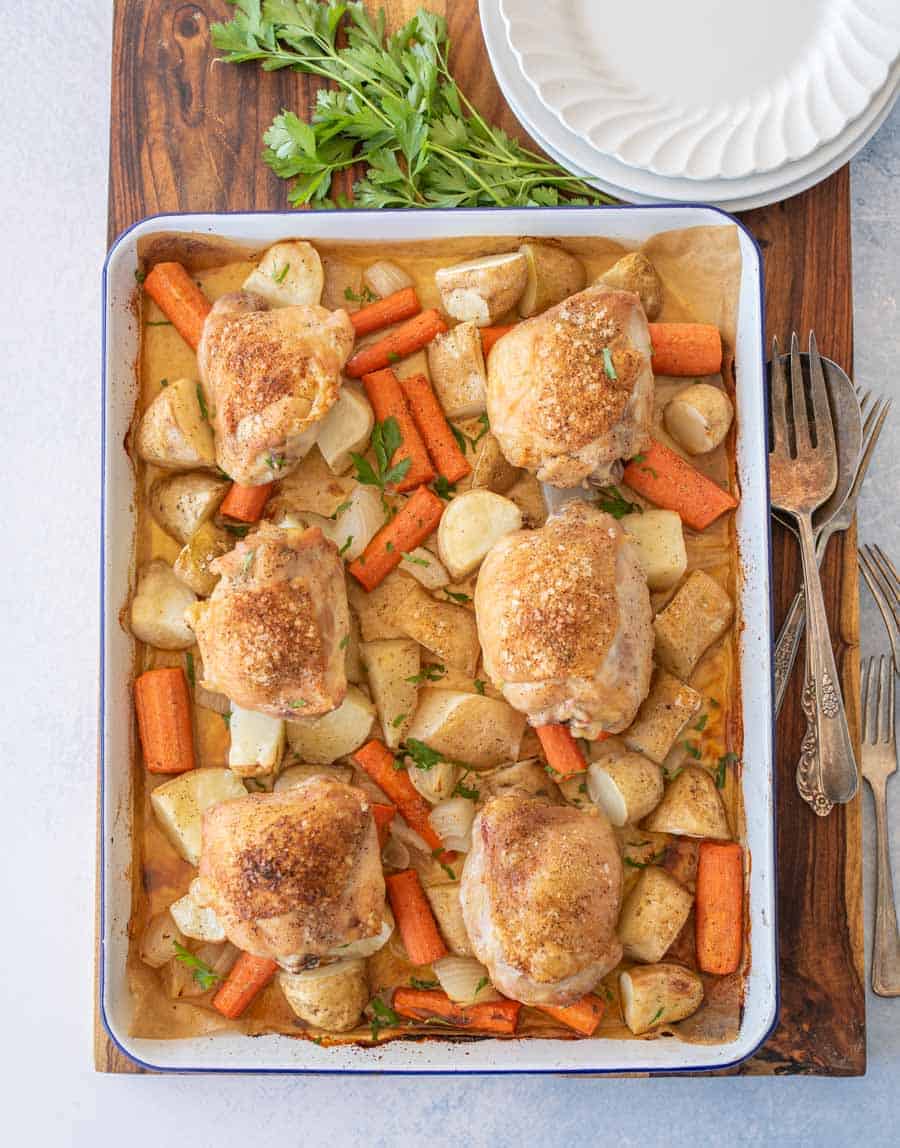 Six oven roasted chicken thighs, carrots, potatoes, and onions on an enamel baking sheet with dinner plates and forks next to it.