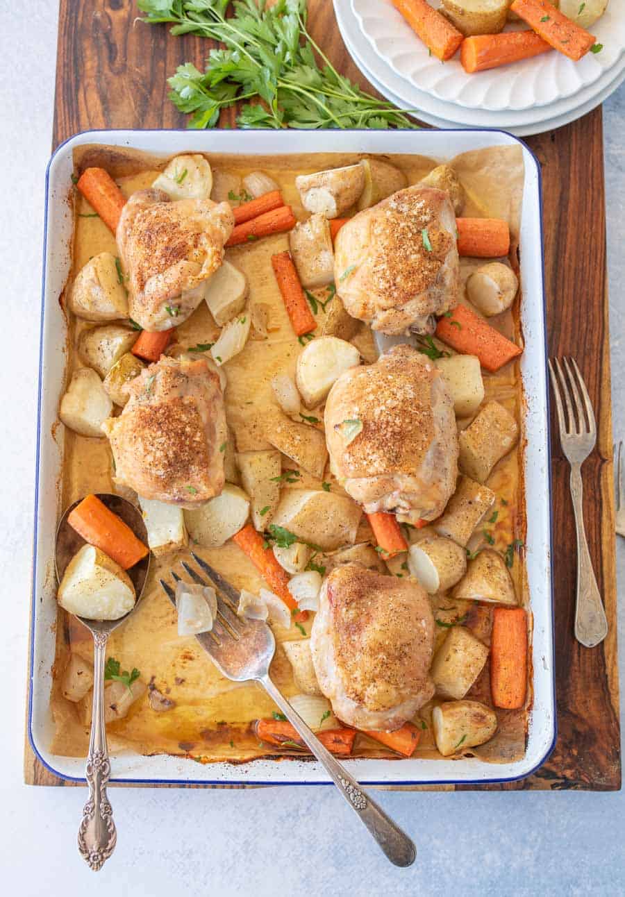 A baked sheet pan meal featuring roasted chicken thighs and potatoes, golden and tender alongside carrots, garnished with fresh herbs. Serving utensils rest in the pan, with a plate of food, fork, and parsley nearby.