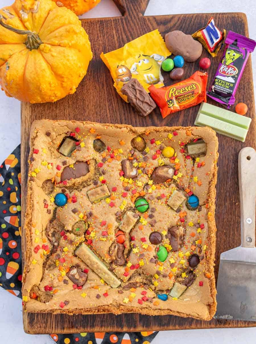 Uncut cookie bars next to a pumpkin and leftover Halloween candy on a wooden cutting board.