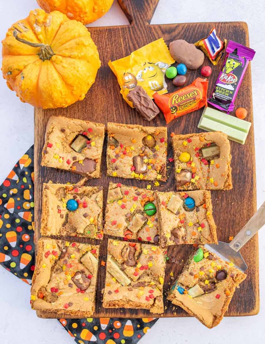 A pan of Halloween candy cookie bars cut up next to a pumpkin and some leftover Halloween candy on a wooden board.