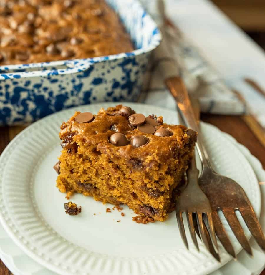 A piece of chocolate chip pumpkin cake on a white plate with a fork and the full pumpkin cake in the background.