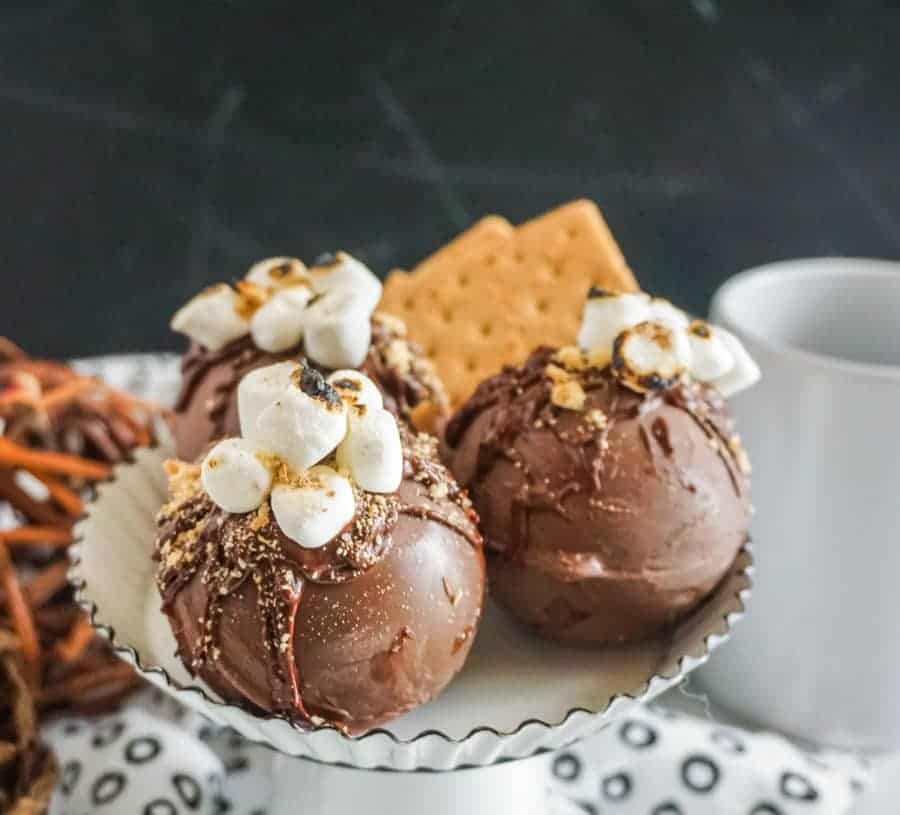 Three finished s'mores hot chocolate bombs on a white enamel cake stand with black rim.