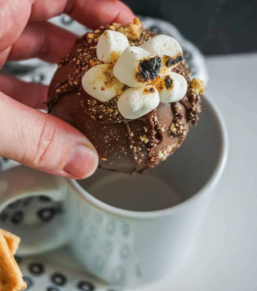 S'mores hot chocolate bomb held in a hand above a white mug.