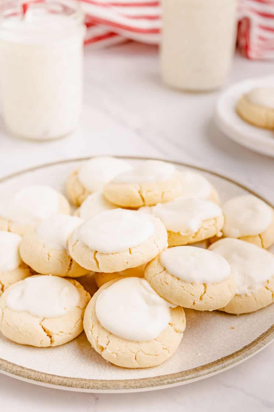 A pile of baked and iced melt away cookies on a cream-colored plate with a glass of milk in the background.