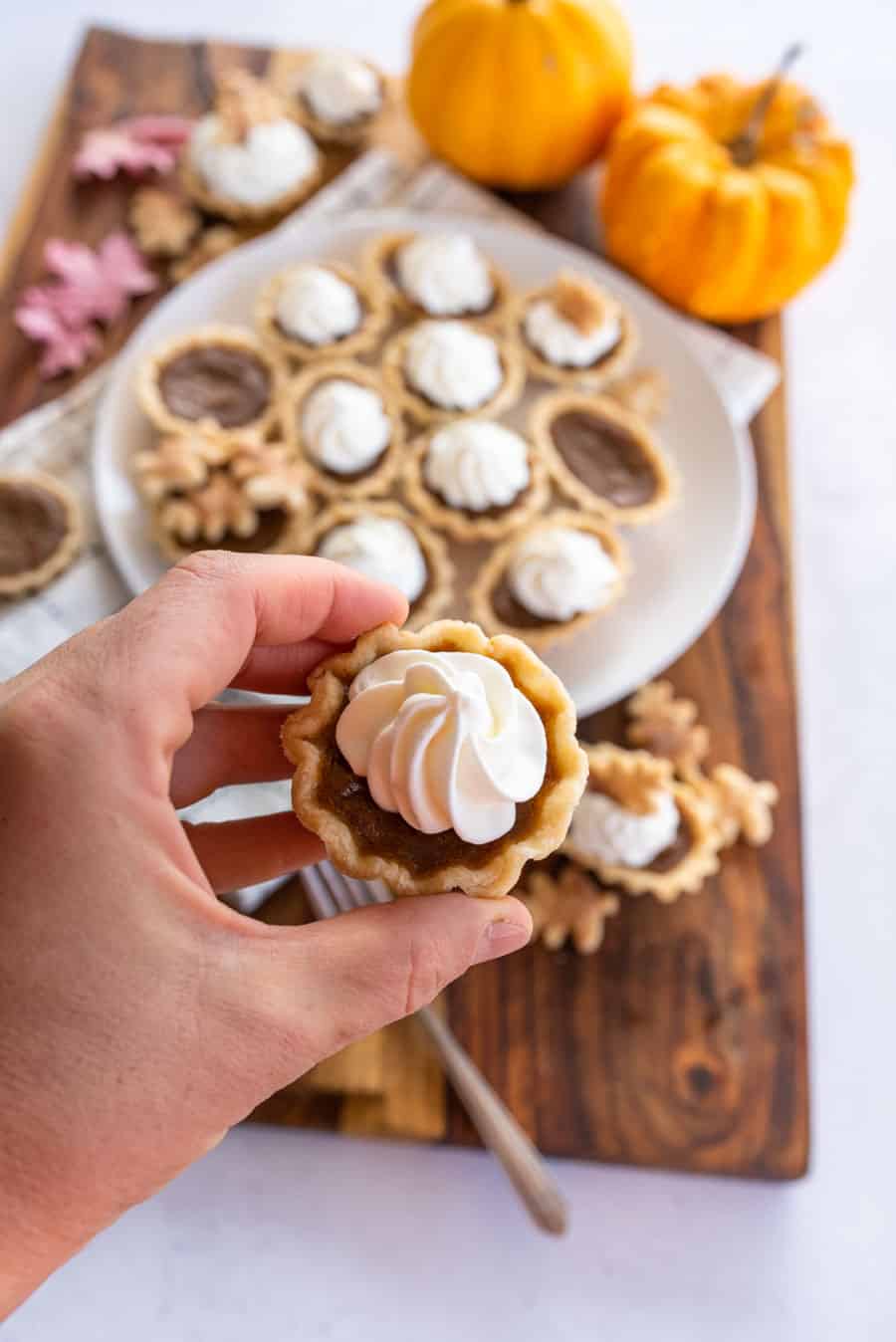 A hand holding a mini pumpkin pie with whipped cream swirled on top with more mini pumpkin pies and a couple pumpkins in the background.