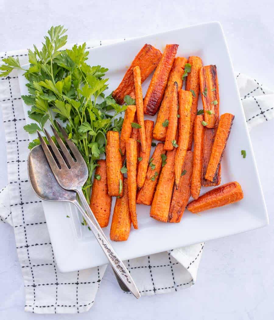 Roasted carrots on a white plate with a fork and spoon with a white and black napkin in the background.