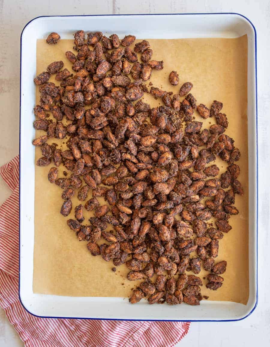 Top view of candied almonds cooling on brown parchment paper on a white baking sheet with a blue rim.