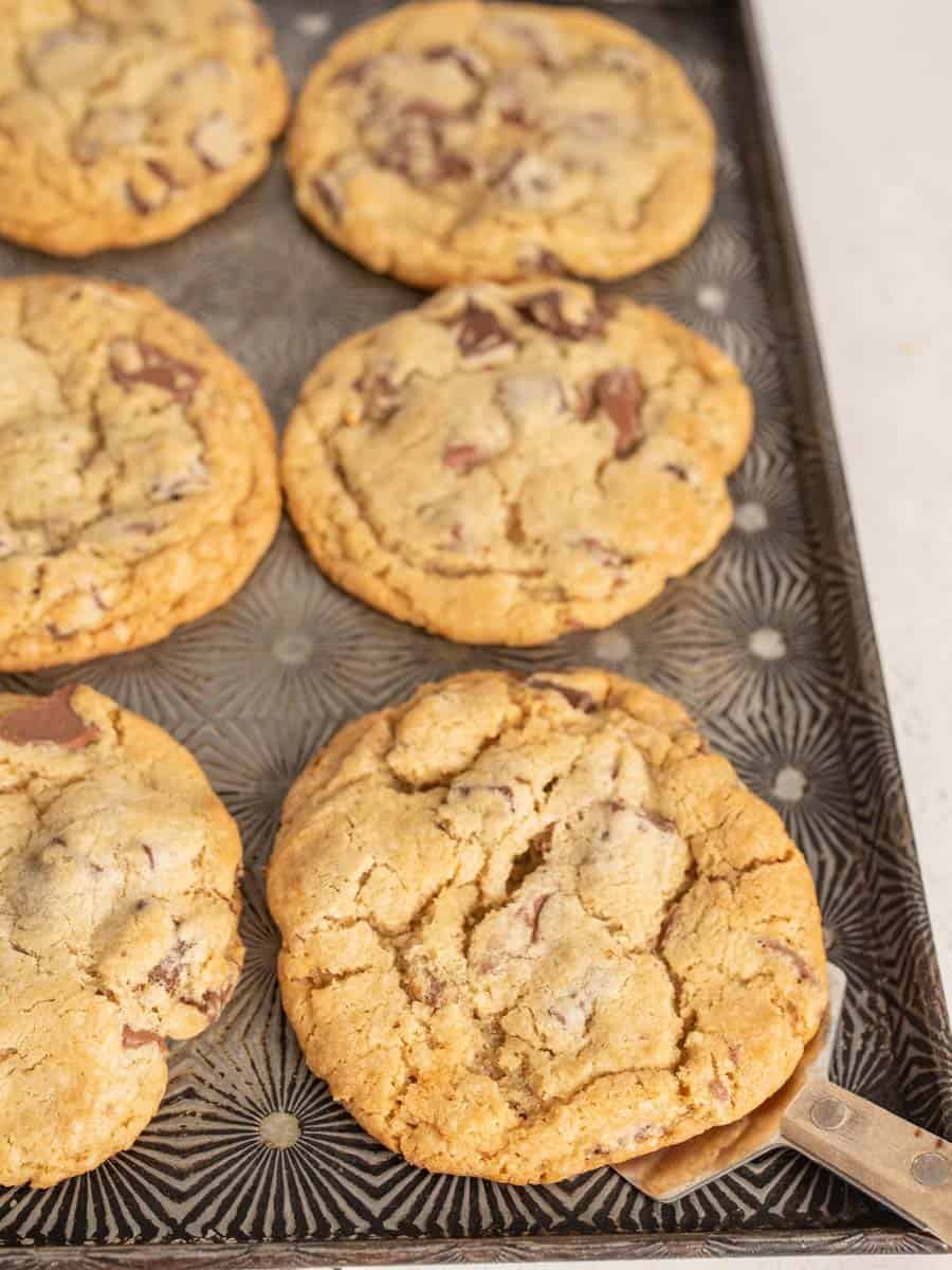 A close up of a thing metal spatula scooping a big chocolate chunk cookie on a baking sheet.