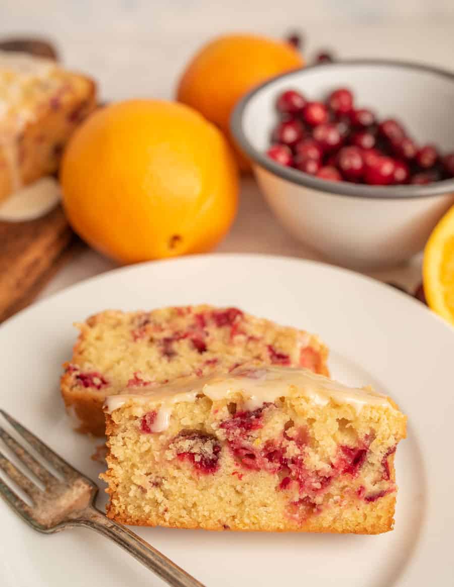 Two slices of cranberry orange bread on a white plate with a fork next to them and with two oranges and a bowl of fresh cranberries in the background.