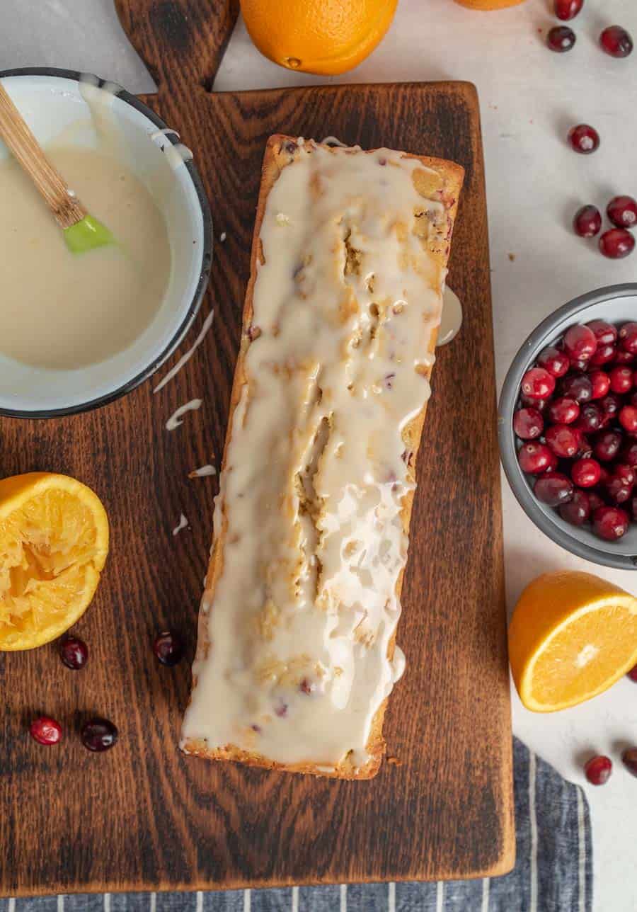 Top view of cranberry orange bread on cutting board with bowl of glaze, bowl of cranberries, and oranges around it.