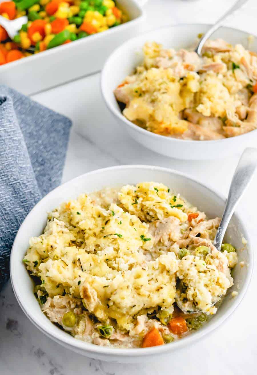 Cooked chicken stew with dumplings in a white bowl with a spoon with a second bowl and some extra vegetables in the background.