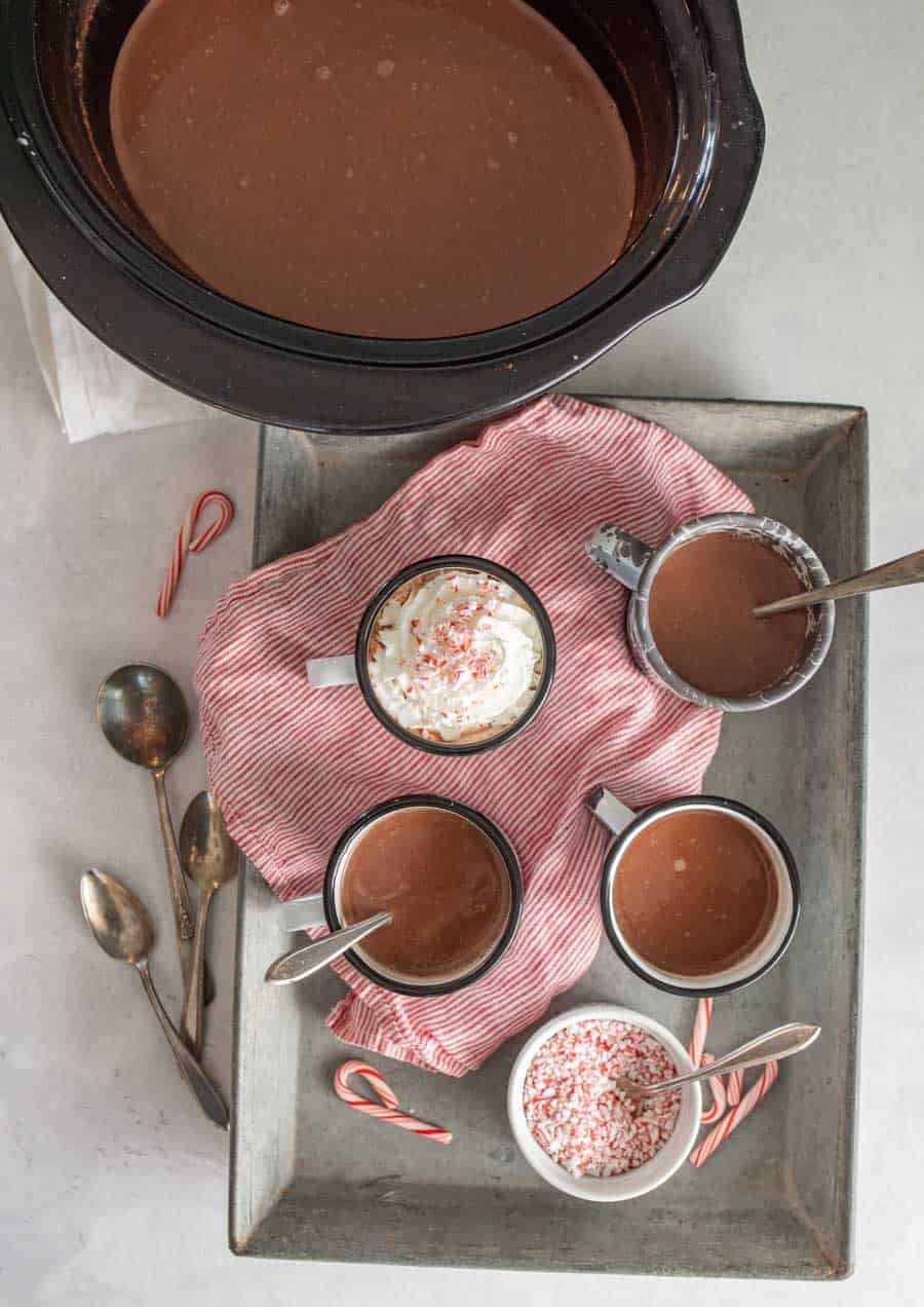 Top view of tray of mugs filled with crock pot hot chocolate next to the crock pot of hot chocolate, some spoons, and crushed candy canes in a ramekin.