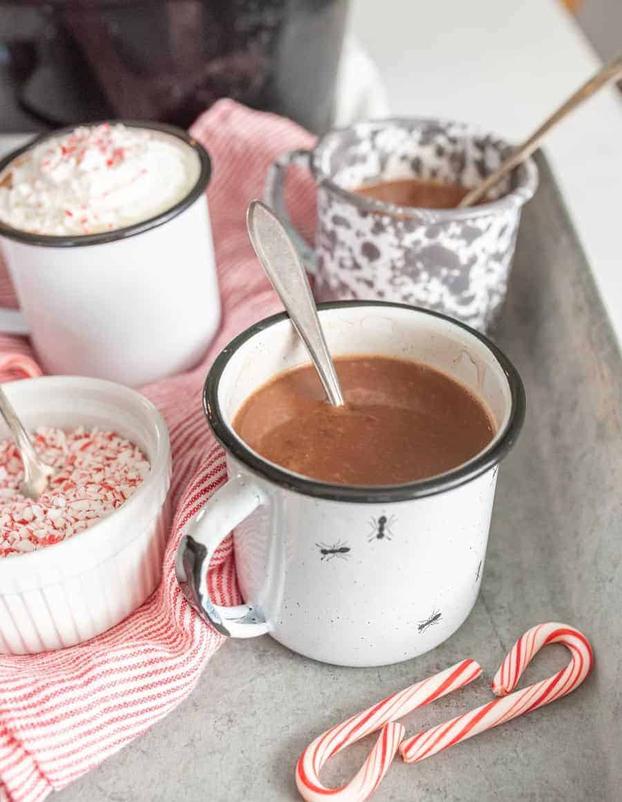 Mugs filled with crock pot hot chocolate with a spoon in them with crushed candy canes in a ramekin next to them.