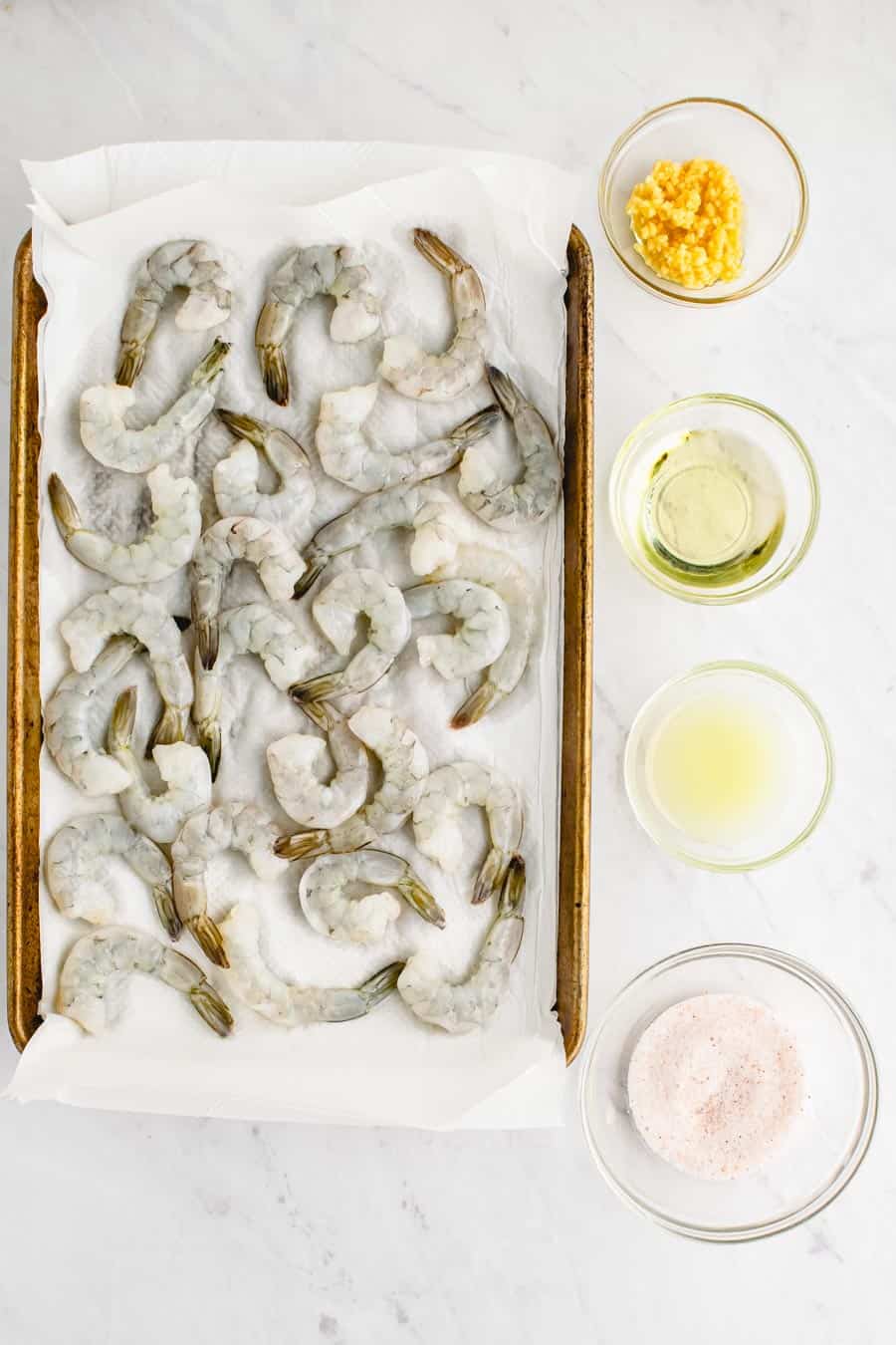 The ingredients to make garlic shrimp including raw shrimp on a paper towel on a baking sheet and some minced garlic, oil, lemon juice, and salt each in a small clear glass bowl.