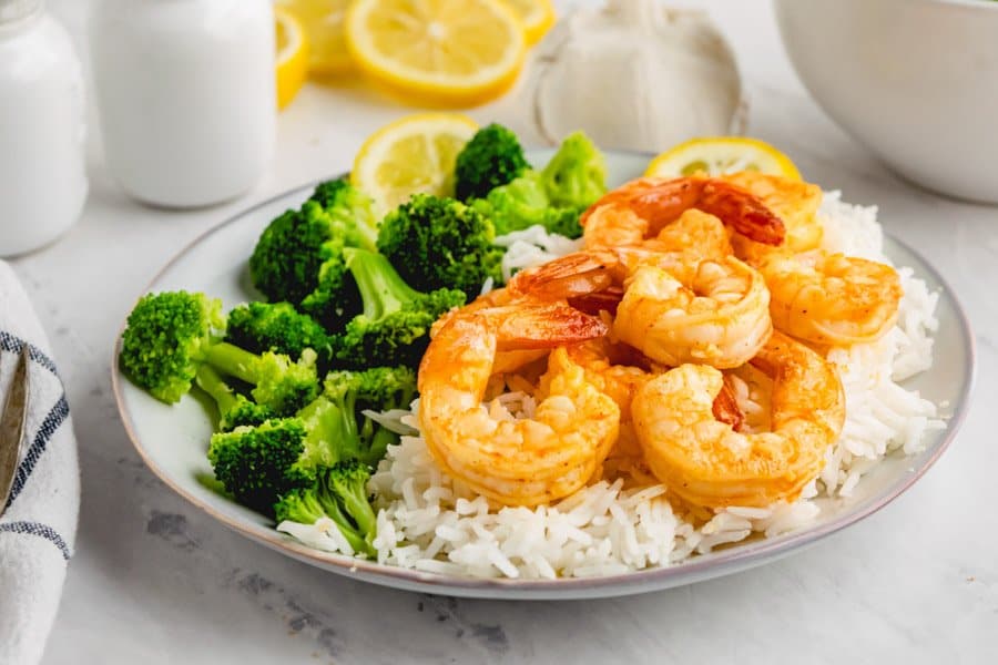 Seven cooked garlic shrimp on top of white rice next to some steamed broccoli florets on a light grey plate.