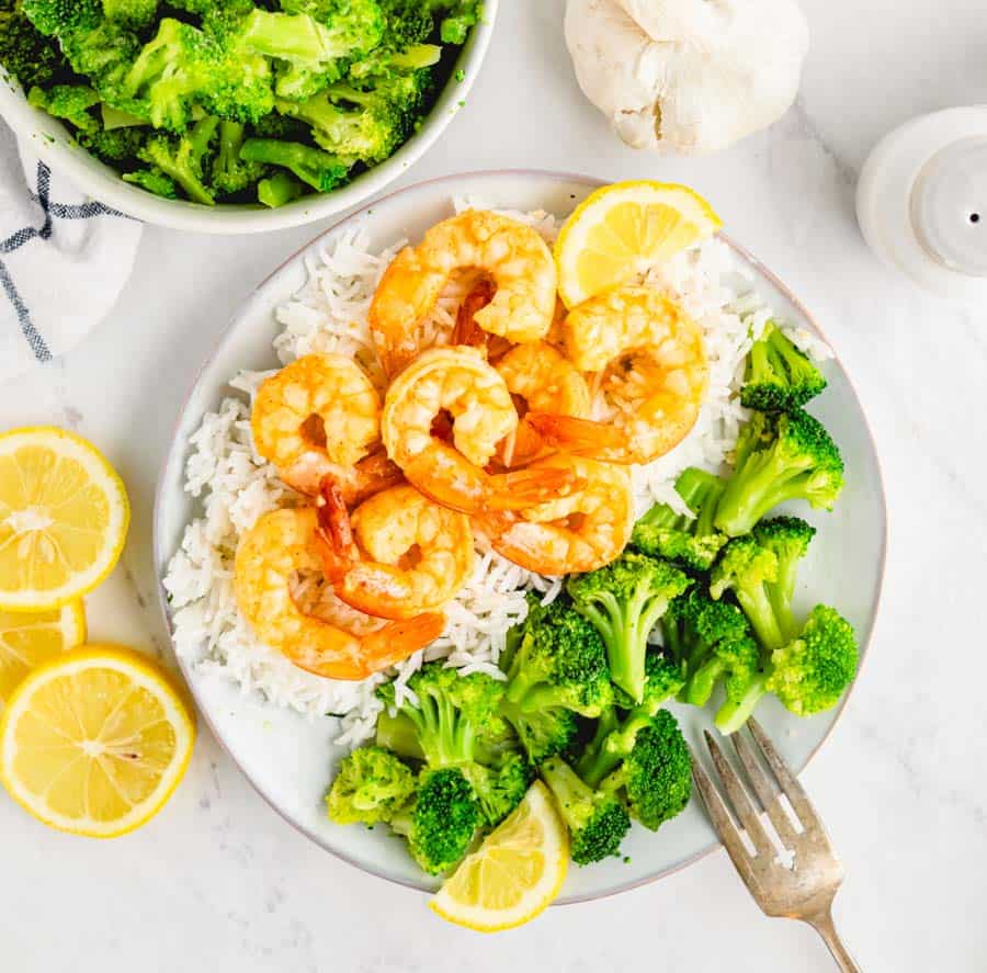 Eight cooked garlic shrimp on top of white rice next to some steamed broccoli florets and there is a bowl of broccoli and a bulb of garlic and some lemon slices in the background.