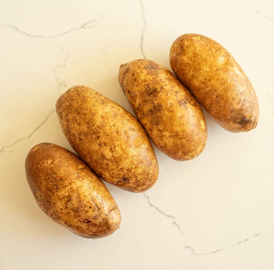 Four unpeeled russet potatoes, perfect for oven baked potatoes, are arranged in a diagonal line on a light-colored, slightly marbled surface.