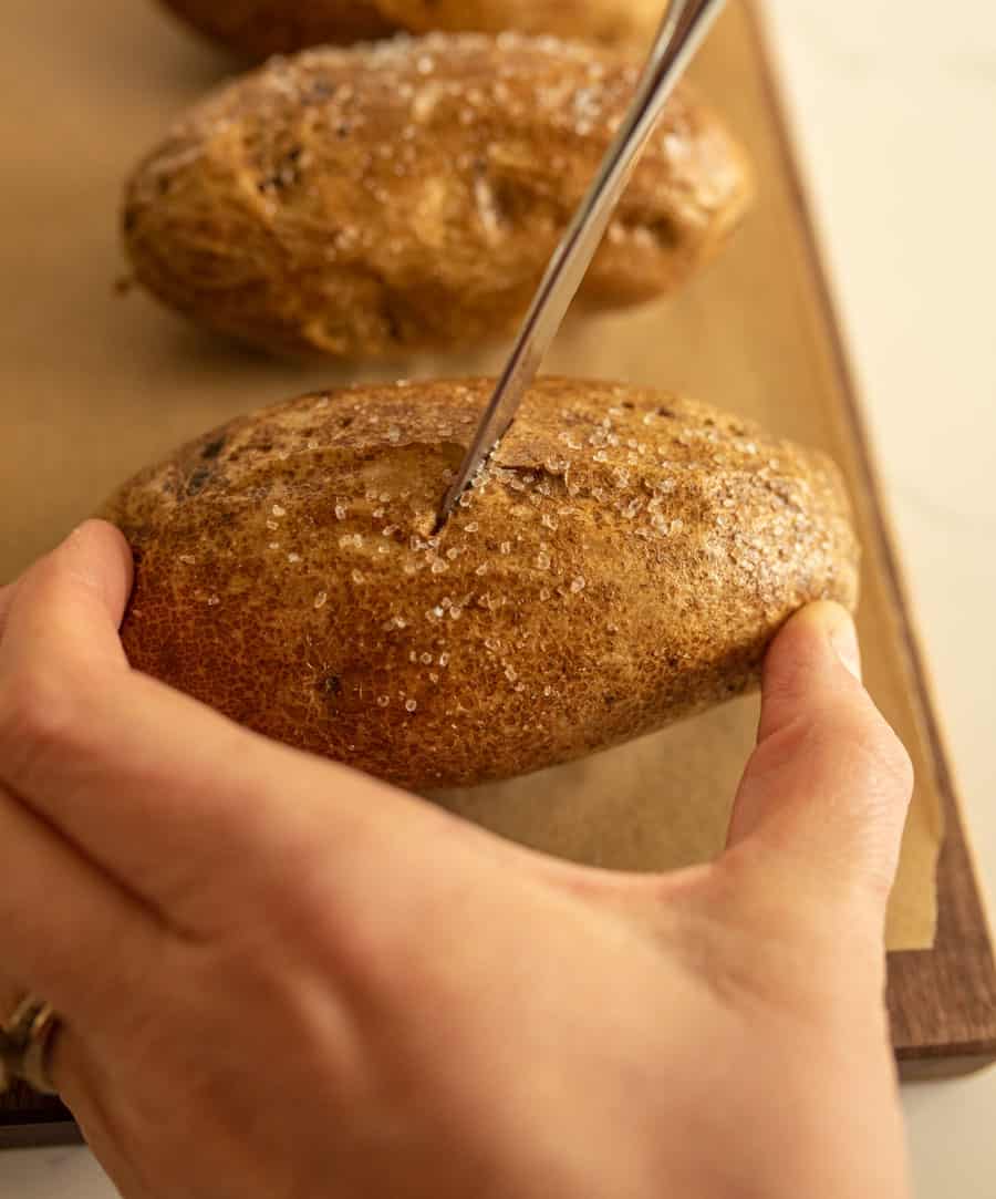A hand holds a salted, unpeeled potato while piercing it with a fork on a parchment-lined baking sheet, preparing oven baked potatoes. Other potatoes are visible in the background.