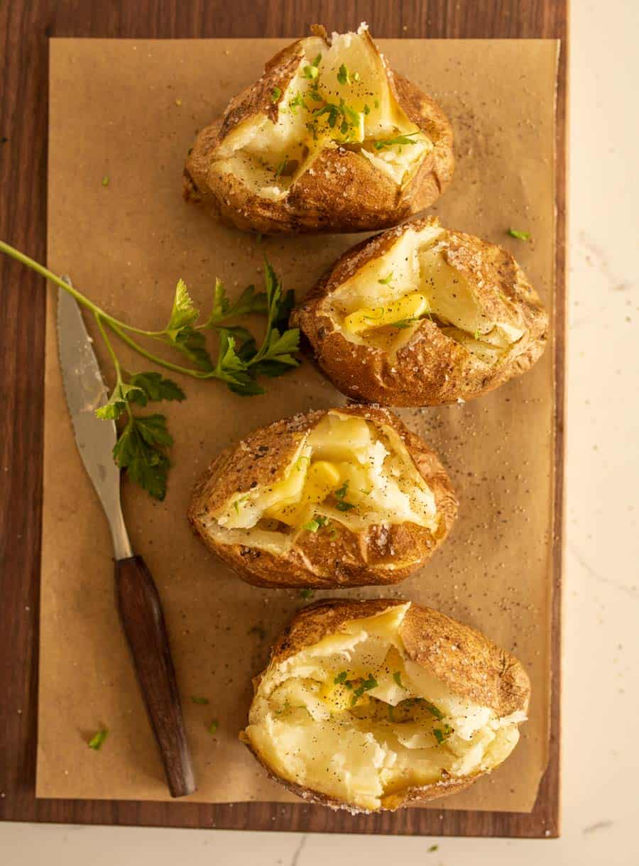 Four oven baked potatoes sliced open with butter melting inside, garnished with chopped herbs, are arranged on parchment paper atop a wooden board. A knife with a wooden handle and a sprig of parsley lie beside them.