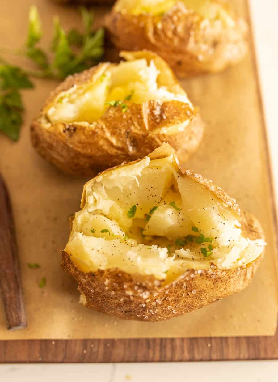 An oven baked potato split open, topped with melting butter, black pepper, and chopped parsley, sits on parchment paper with more oven baked potatoes and fresh parsley in the background.