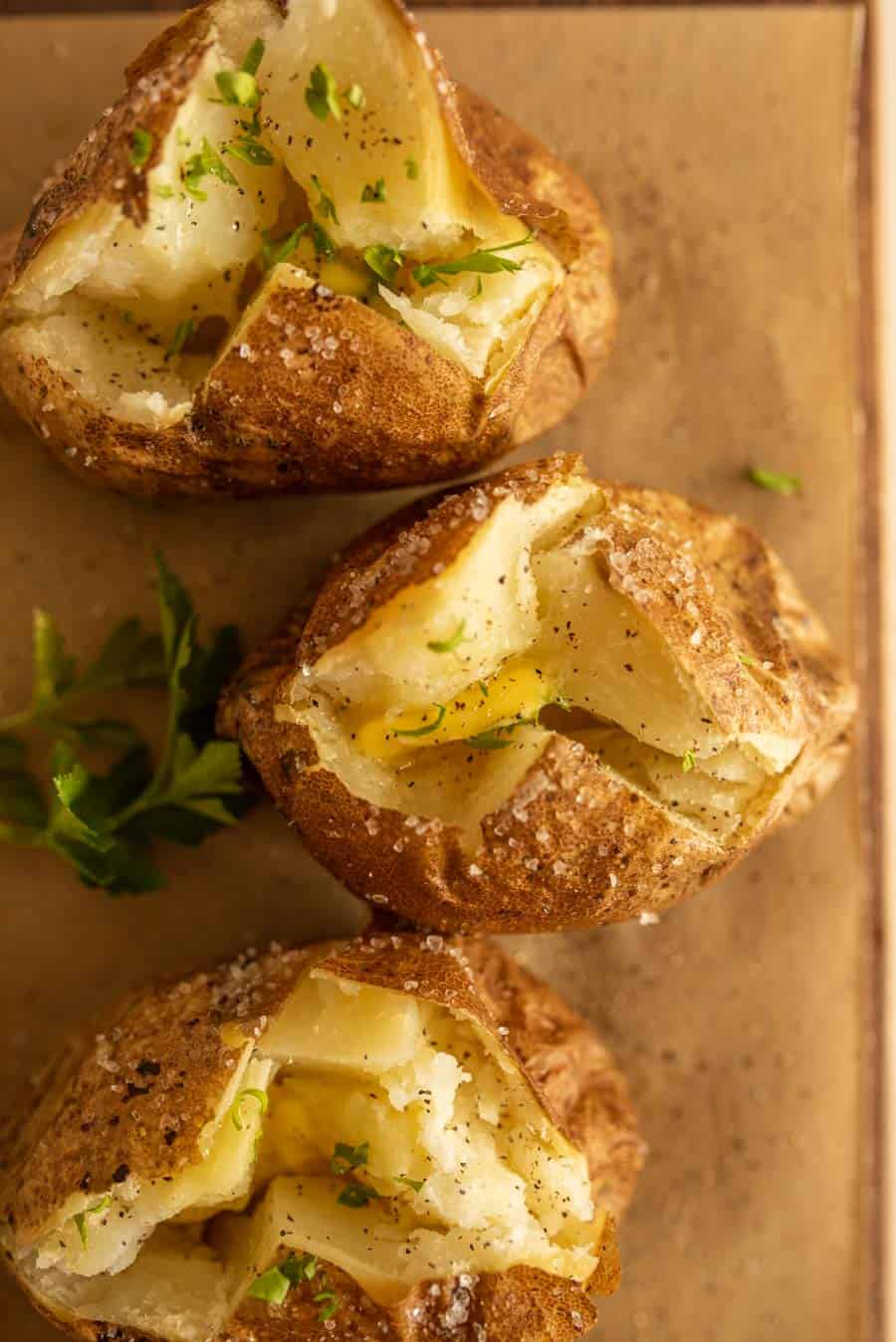 Three oven baked potatoes with crispy skins are split open, revealing fluffy insides topped with melted butter, black pepper, and chopped parsley, resting on a brown surface with a garnish of fresh herbs nearby.