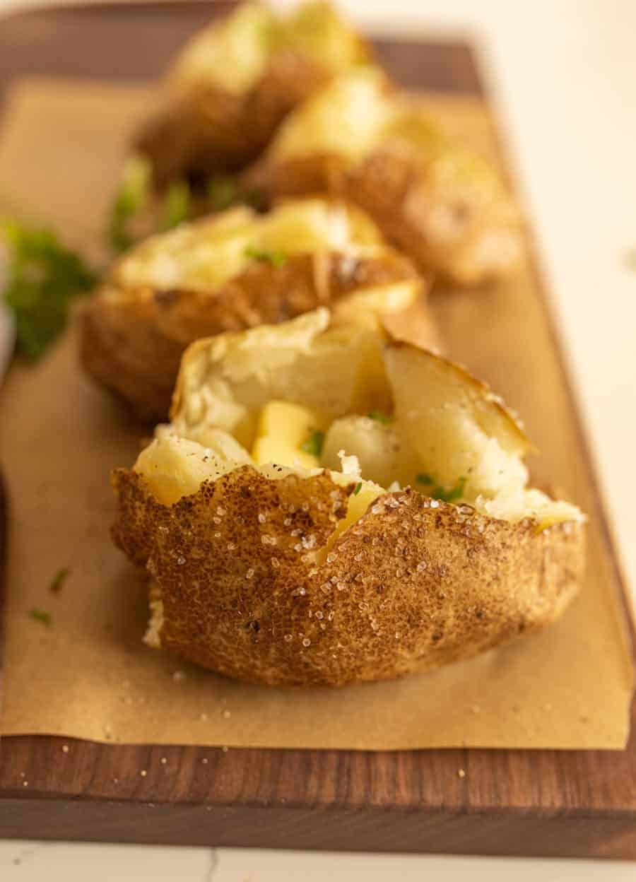 A close-up of oven baked potatoes with crispy, salted skin, split open and topped with butter and a sprinkle of herbs, served on parchment paper on a wooden board. Other potatoes are blurred in the background.