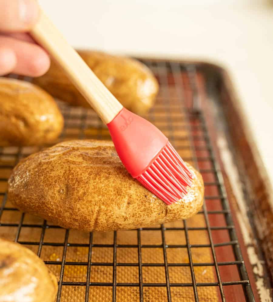 A hand uses a red silicone brush to apply oil to a potato resting on a wire rack over a baking sheet, preparing delicious oven baked potatoes. Other potatoes are visible in the background.
