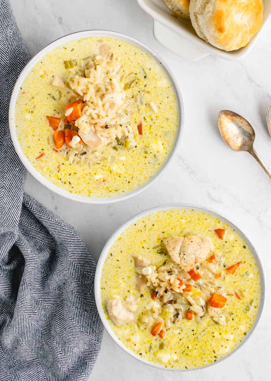 Two bowls of chicken and wild rice soup in white bowls on a white counter with biscuits in a white serving dish in the background.